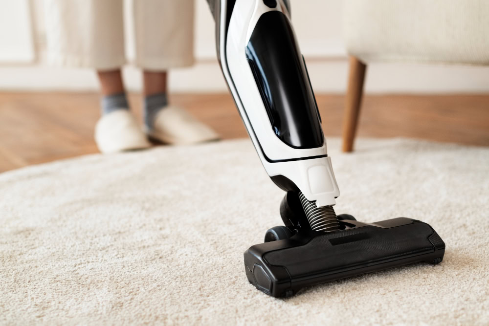 Close-up of a cordless vacuum cleaner being used on a soft, light-colored carpet, with a person wearing slippers visible in the background.