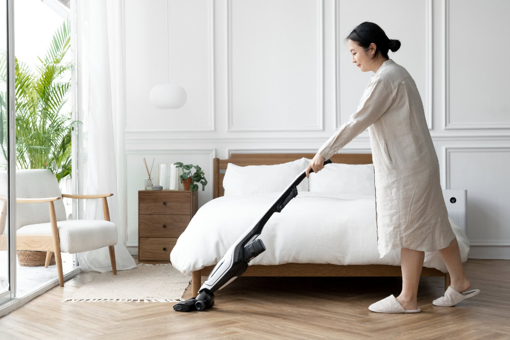 A woman using a cordless vacuum cleaner to clean the wooden floor in a bright and minimalist bedroom with a neatly made bed and indoor plants.