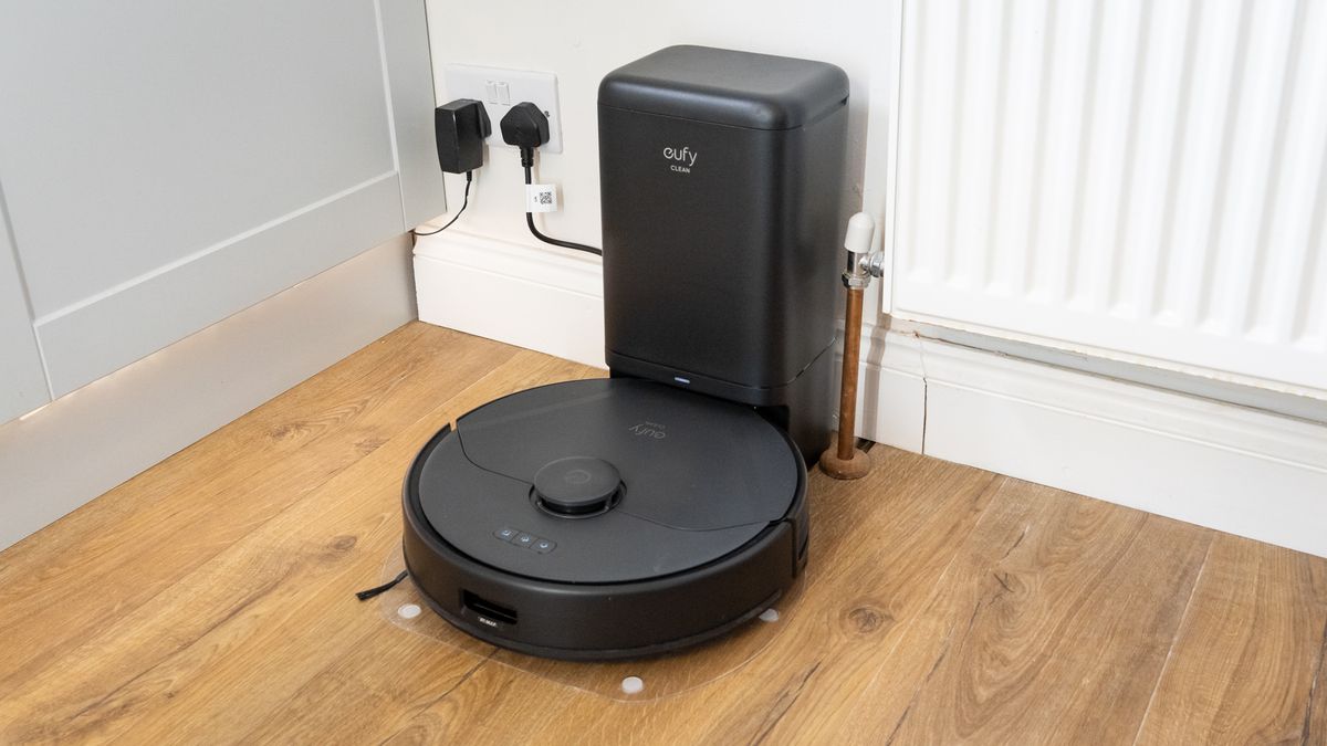 This image shows a robotic vacuum cleaner with a dock or self-emptying station placed in a home setting, near a wall socket and a radiator. Let me know how I can assist you further with this!