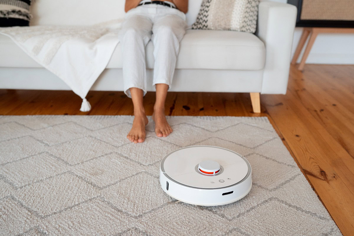 A robotic vacuum cleaner operating on a textured rug in a living room, with a person sitting on a white sofa in the background.