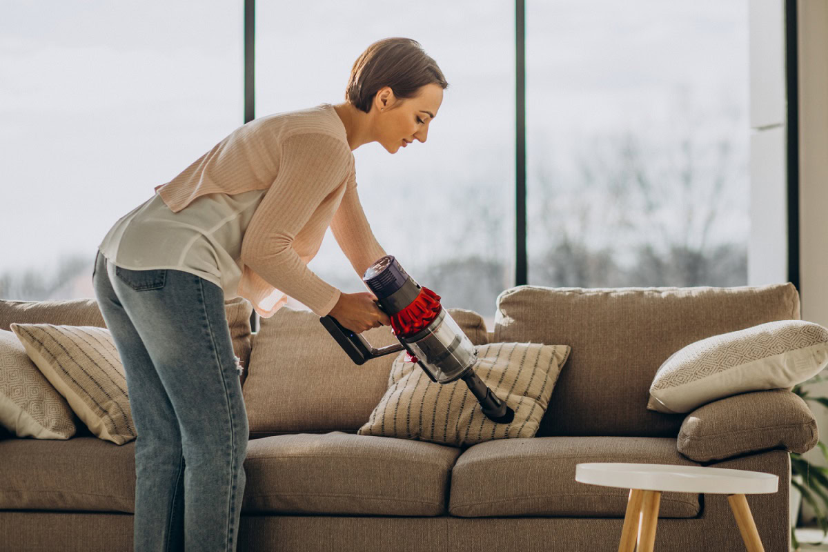 A woman using a cordless vacuum cleaner to clean a beige sofa in a bright living room with large windows in the background.