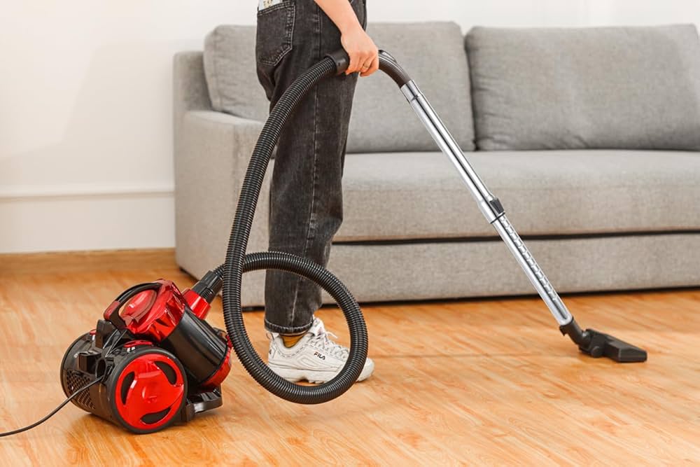 Person using a red canister vacuum cleaner to clean a wooden floor, with a gray sofa in the background.