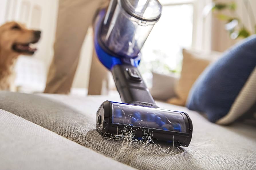 Close-up of a cordless vacuum cleaner with a motorized brush attachment removing pet hair from a fabric surface, with a golden retriever in the background.