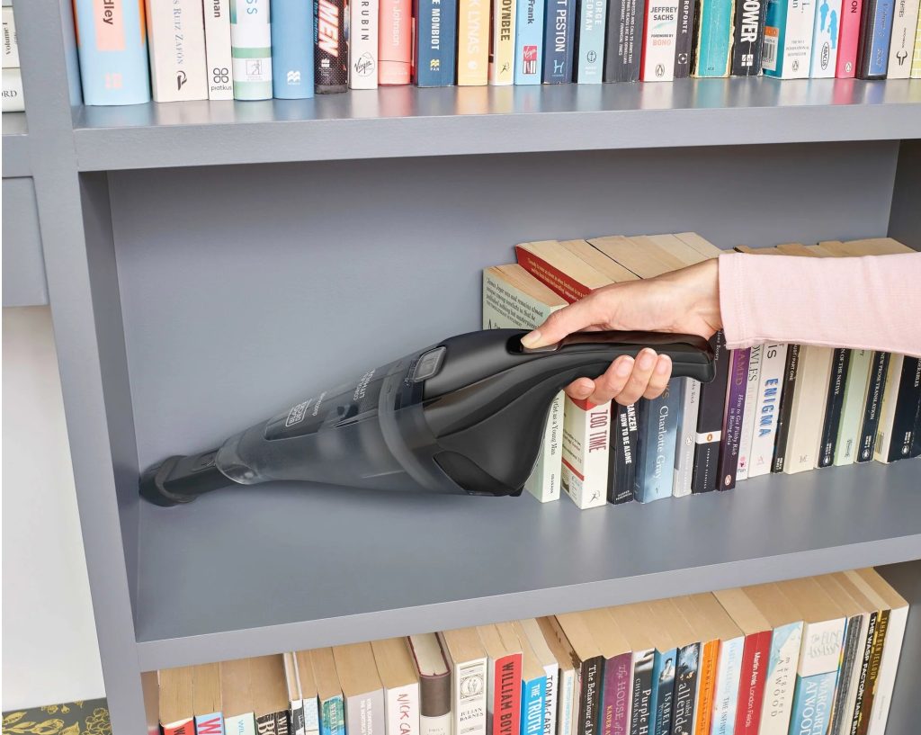 Person using a handheld vacuum cleaner to clean dust from a bookshelf filled with colorful books.