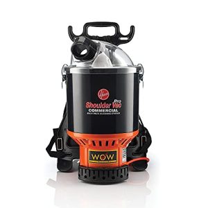 The image shows a Hoover Shoulder Vac Pro Commercial backpack vacuum cleaner. The design is compact and ergonomic, featuring a black and orange body with the branding "Shoulder Vac Pro" and "Commercial" prominently displayed. It includes adjustable straps for wearing on the back and a sturdy handle on top for easy portability. This vacuum cleaner is intended for heavy-duty or professional cleaning tasks and offers efficient suction power for commercial environments.