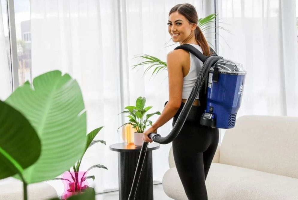 The image depicts a person vacuuming an office space using a backpack vacuum cleaner. They are wearing the vacuum cleaner on their back, and it appears compact and ergonomic. The person is holding the vacuum wand and cleaning a carpeted floor in a bright office environment. The office has modern furniture, such as chairs and desks, with natural light streaming through the windows.