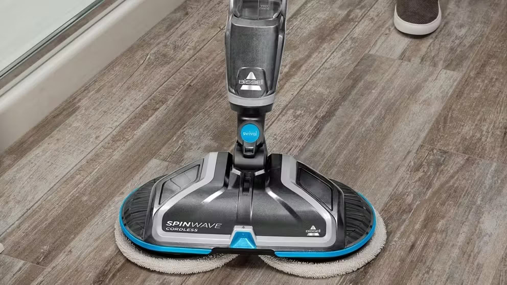 Close-up of a Bissell SpinWave cordless floor cleaner with dual spinning pads, shown on a wooden floor surface.