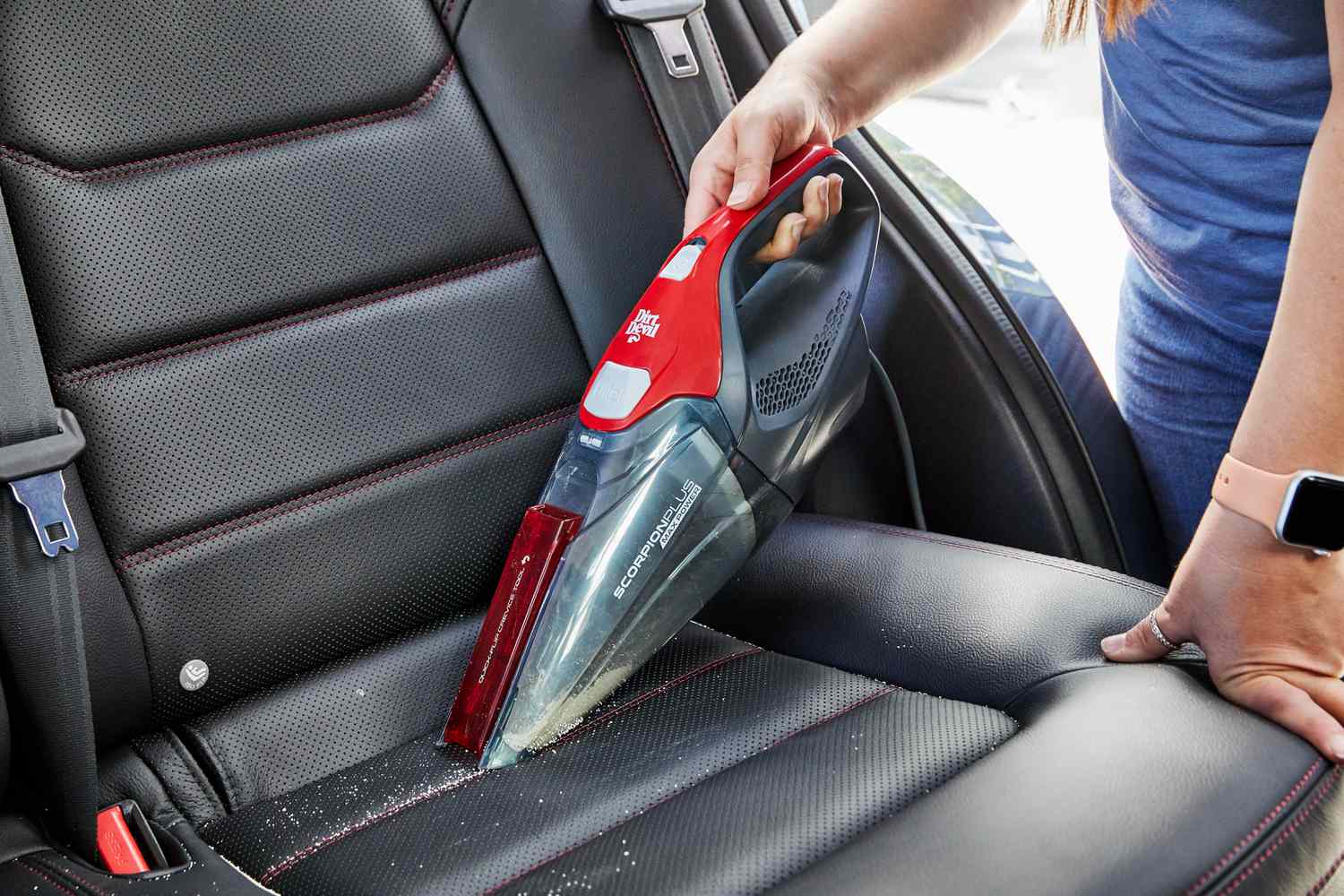 Close-up of a person using a red handheld vacuum cleaner to remove debris from the leather seat of a car interior.