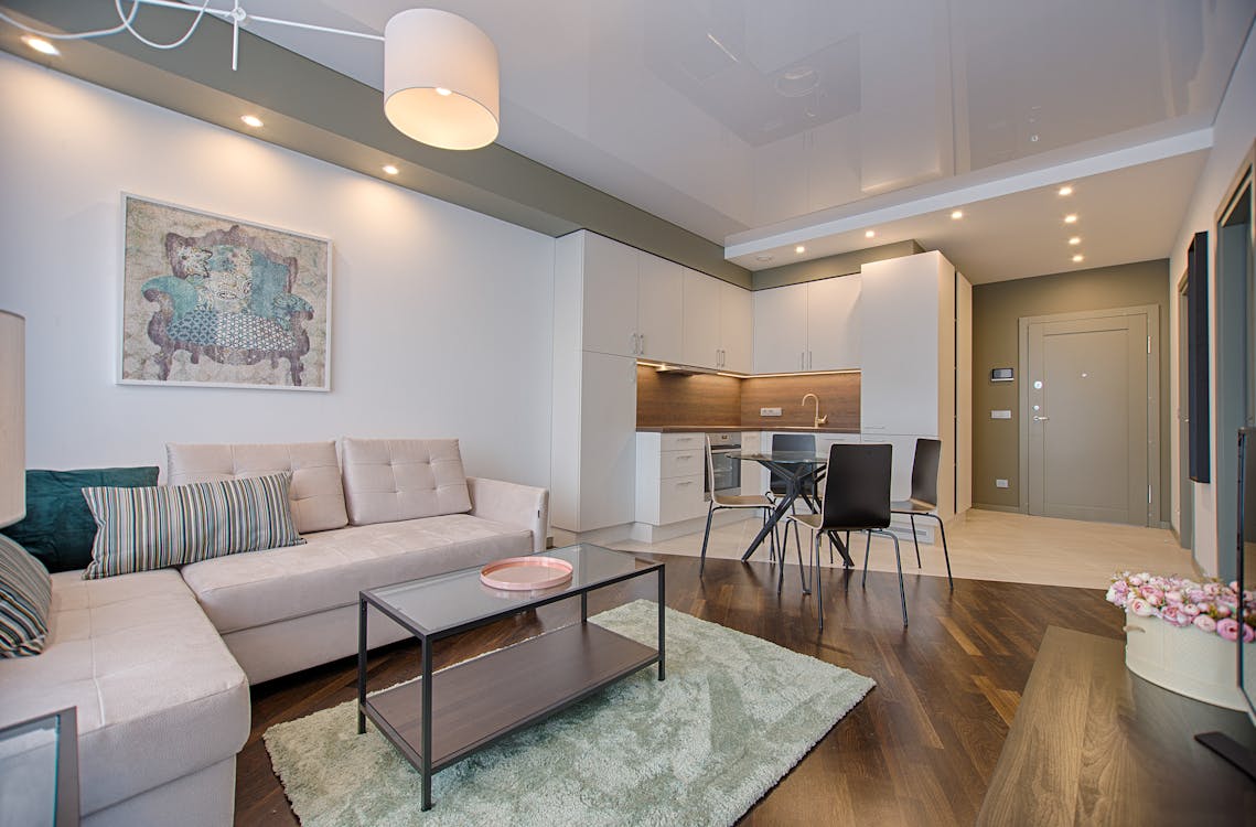 Modern living room with a beige sofa, striped cushions, a glass coffee table on a green rug, and an open kitchen in the background featuring white cabinets and warm wooden accents.