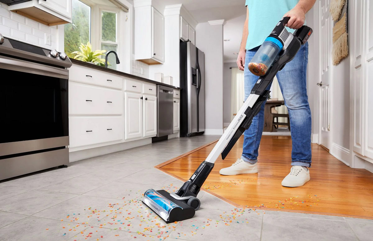 Person using a cordless vacuum cleaner to clean spilled cereal from a tiled and wooden kitchen floor, with modern white cabinetry and appliances in the background.