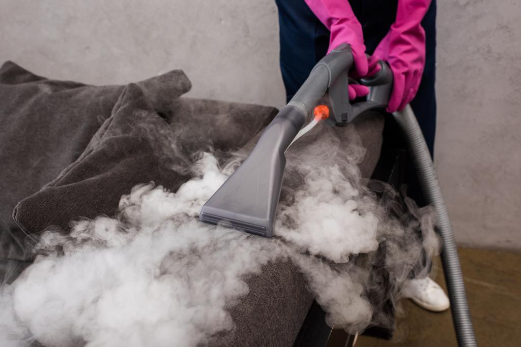 Close-up of a person wearing pink rubber gloves using a steam vacuum cleaner to sanitize a dark fabric surface, with visible steam rising during the cleaning process.