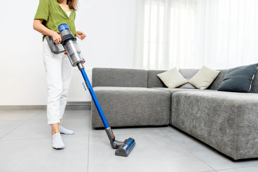 Person using a cordless vacuum cleaner with a blue handle to clean a tiled floor in a bright living room, featuring a gray sectional sofa with decorative cushions.